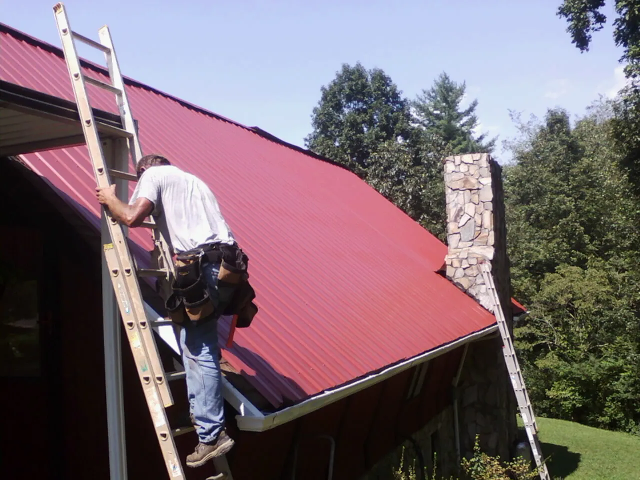 Roofer on a ladder beside a red metal roof and stone chimney — Soffit & Fascia Repair by Roofing Brothers in Bates City